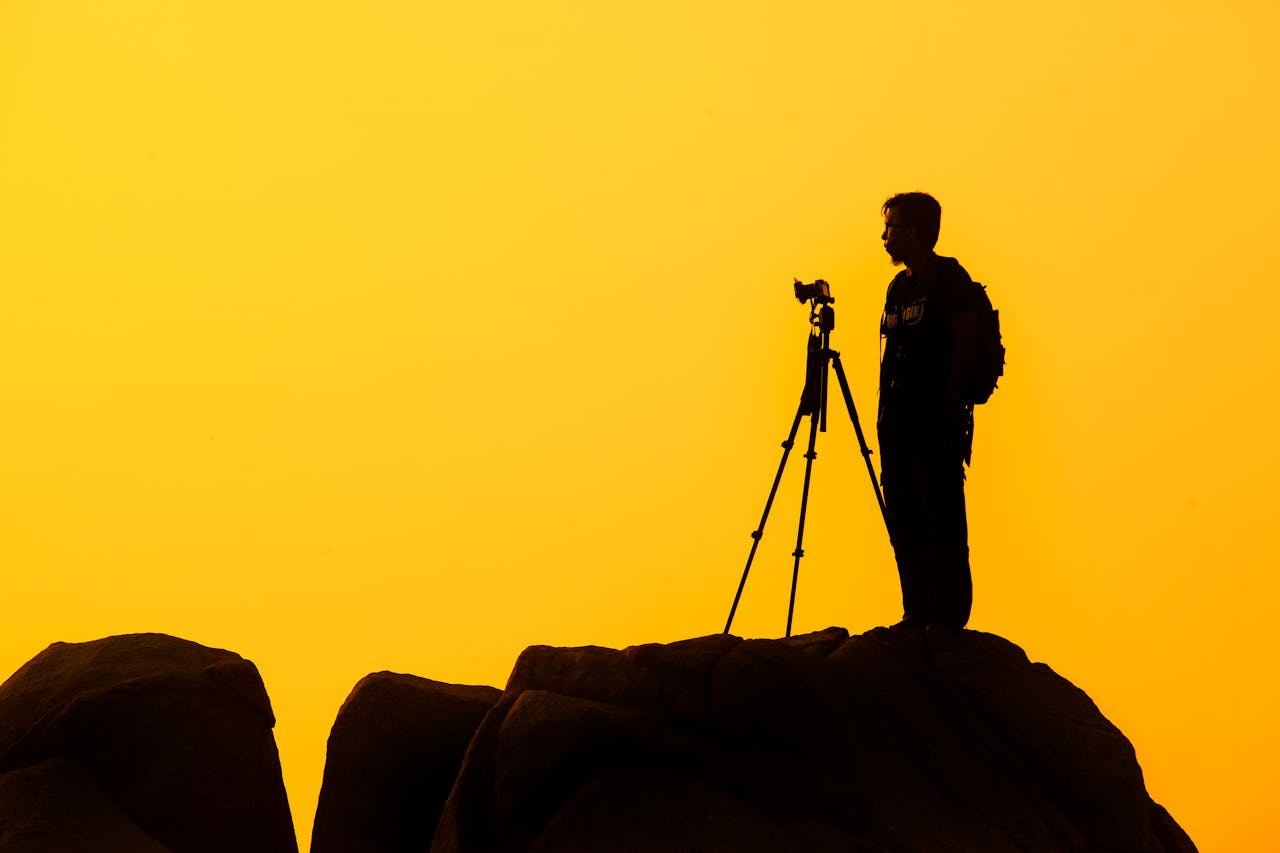 hero-img-01 Silhouette of a lone photographer with tripod on a rocky cliff against a golden sunset sky.